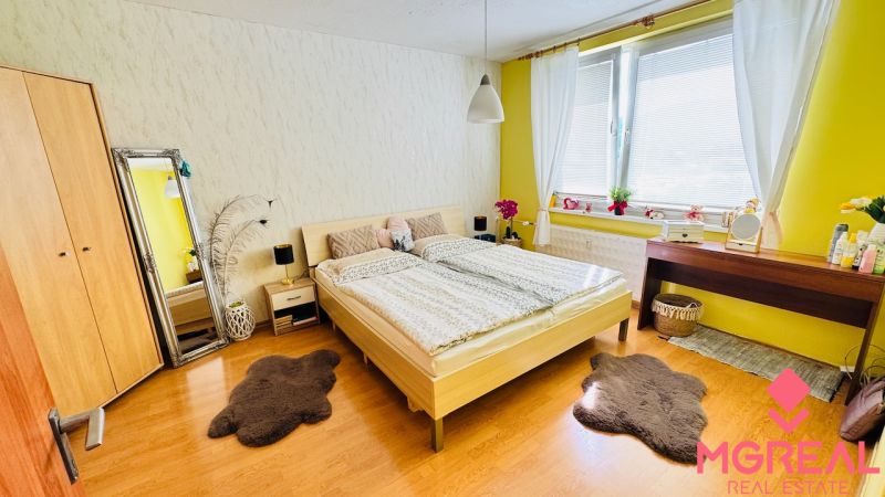 Schlafzimmer in einer Einzimmerwohnung mit Holzdekorboden, hellen Wänden, Fenstern mit weißen Vorhängen.