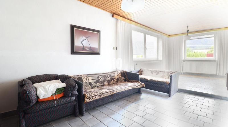 Ein Zimmer in einem Einfamilienhaus mit einem Sofa, einem Bild an der Wand und großen Fenstern.