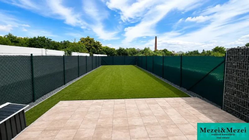 Der Garten eines Einfamilienhauses in Trnava mit Rasen und einer gepflasterten Terrasse unter blauem Himmel.