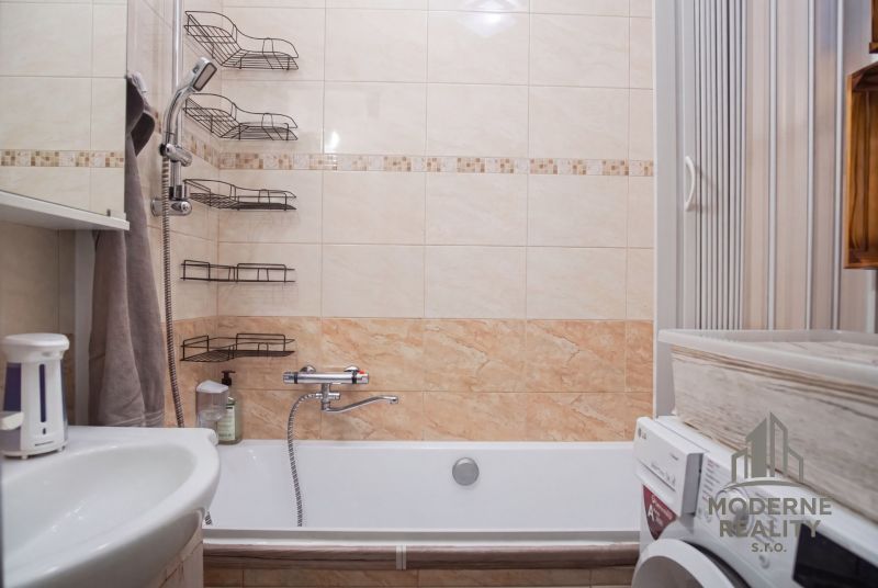 Badezimmer mit Badewanne und Waschmaschine in einer 2-Zimmer-Wohnung. Fliesen mit beigem Dekor.