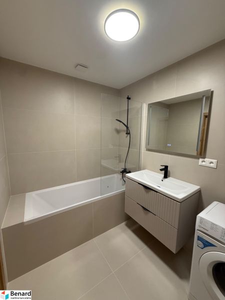Badezimmer in einer 3-Zimmer-Wohnung mit Badewanne, Waschbecken und Waschmaschine, beige Fliesen.