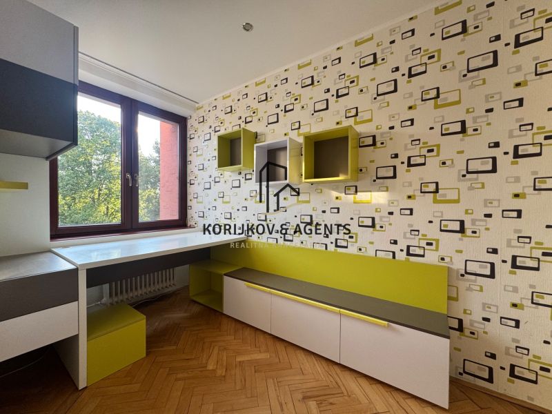 Ein Zimmer in einer 4-Zimmer-Wohnung mit geometrischer Tapete, Möbeln und einem Boden in Holzoptik.