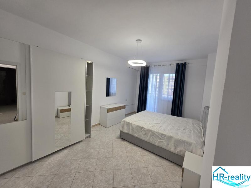 Helles Schlafzimmer mit Bett, Schrank, Fliesen und Vorhängen in einer 2-Zimmer-Wohnung.