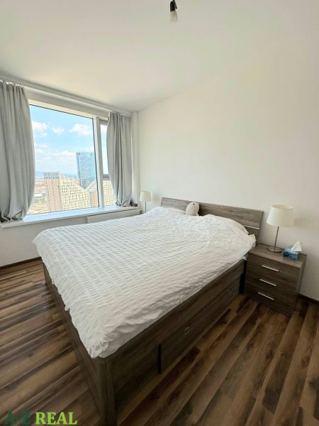 Ein Zimmer mit einem großen Fenster, einem Bett und einem Boden mit Holzdekor in einer 3-Zimmer-Wohnung.