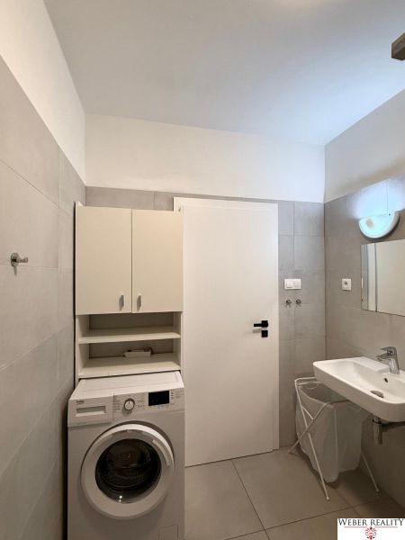 Badezimmer in einer 2-Zimmer-Wohnung mit Waschmaschine, weißem Schrank und Waschbecken auf hellen Fliesen.