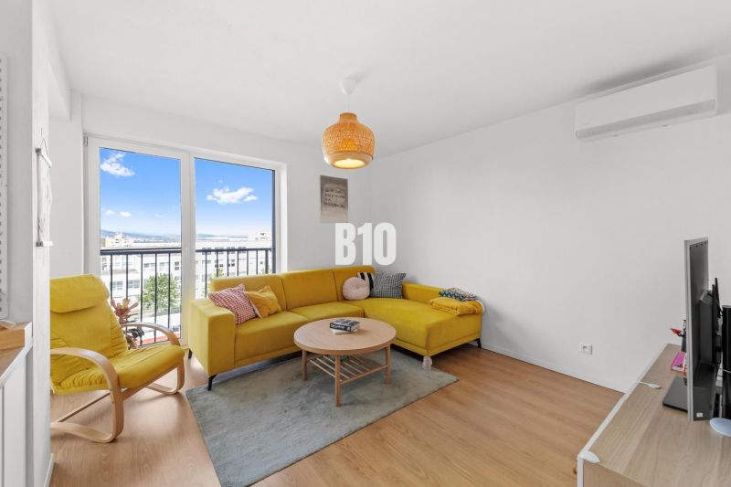 Wohnzimmer einer Vierzimmerwohnung mit gelbem Sofa und großem Fenster, Boden mit Holzdekor.