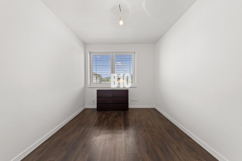 Ein Zimmer in einem Einfamilienhaus mit einem Fenster, einer Kommode und einem Boden in Holzoptik.