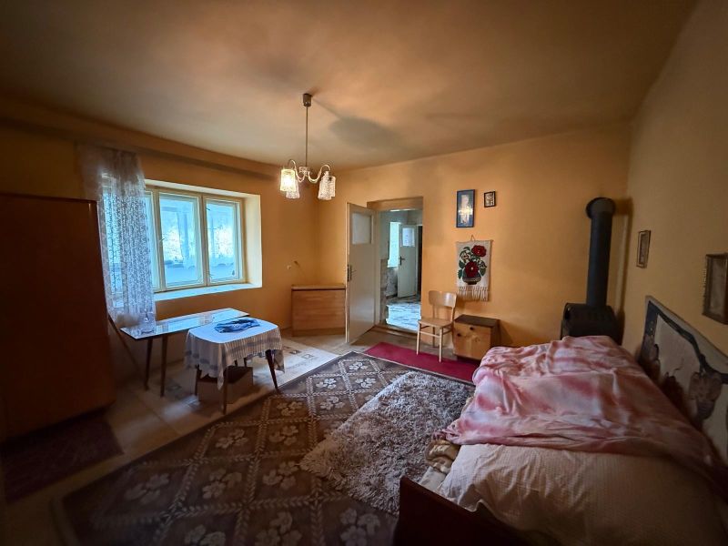 Innenraum eines Einfamilienhauses mit Bett, Tisch und Holzofen, in warmen Farbtönen gehalten.
