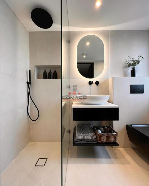 Ein Badezimmer in einem Einfamilienhaus mit beleuchtetem Spiegel, schwarzen Accessoires und einer Dusche.