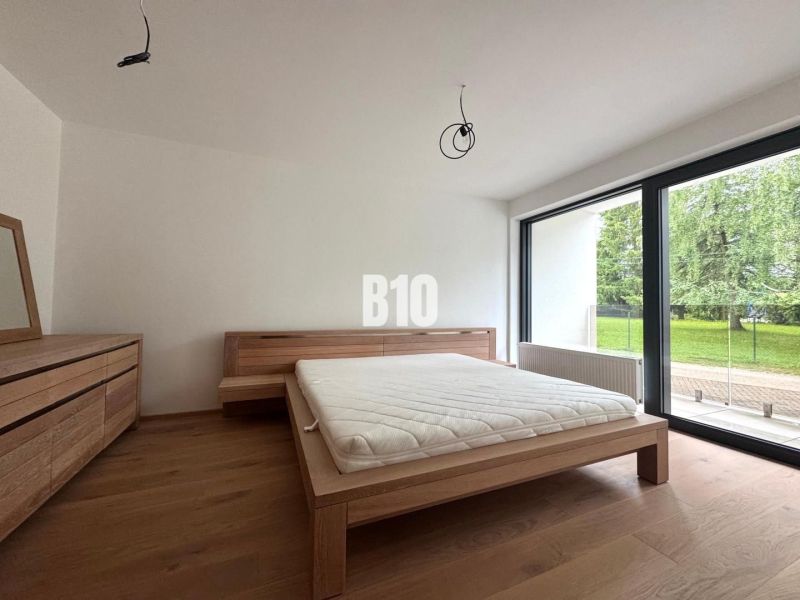 Schlafzimmer mit Bett und Kommode, Boden mit Holzdekor, großes Fenster mit Blick auf den Garten.