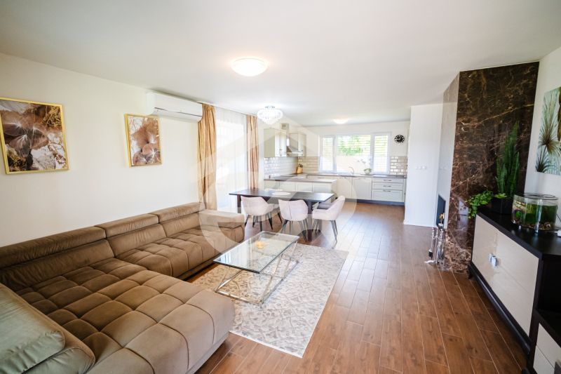 Wohnzimmer in einem Einfamilienhaus mit Küche, beigem Sofa und Boden in Holzoptik.