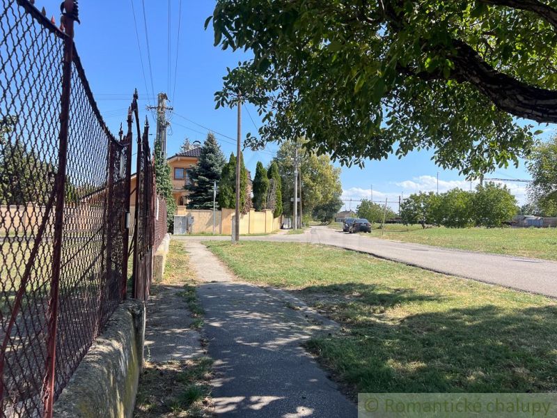Der Gehweg verläuft entlang des Zauns in den Gärten von Bernolákovo, mit der Straße im Hintergrund.
