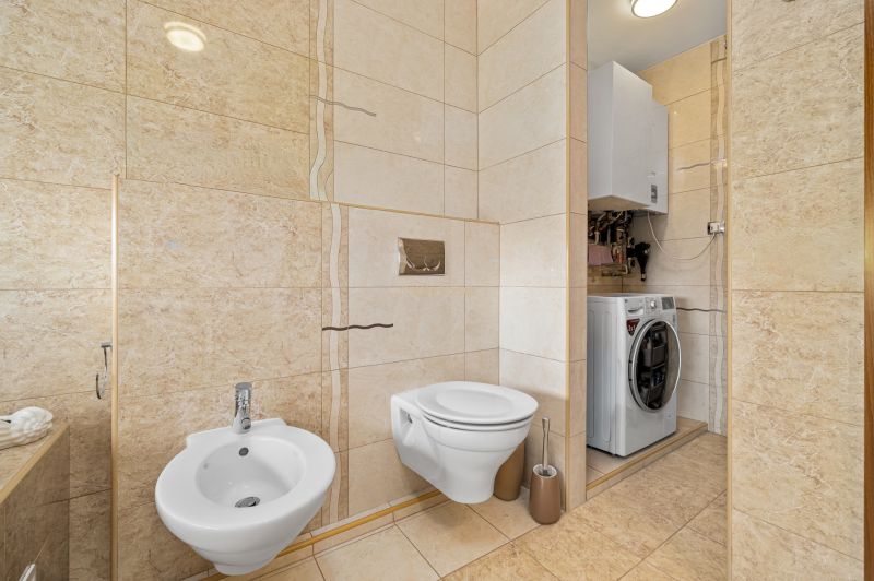 Badezimmer mit Toilette, Bidet und Waschmaschine in einem Einfamilienhaus. Cremefarbene Fliesen und Bodenbelag.