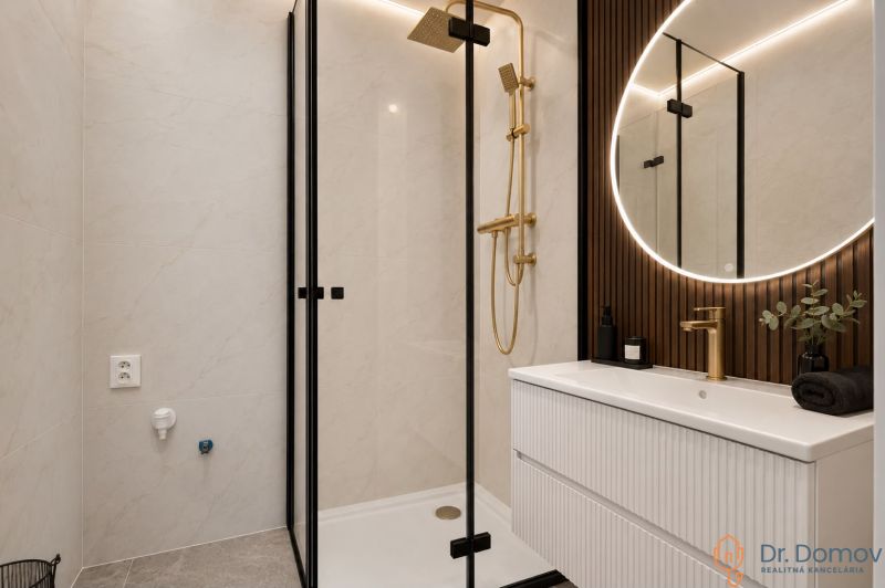 Badezimmer in einer 3-Zimmer-Wohnung mit eleganter Dusche, goldenen Details und beleuchtetem Spiegel.