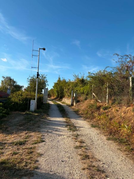 Ein befestigter Feldweg, der zur Hütte in Banka führt, gesäumt von Büschen und einem Zaun.