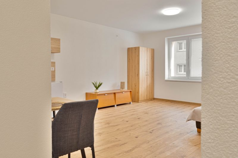 Einzimmerwohnung mit Holzboden, Schrank, Fenster, Tisch und zwei Schränkchen.