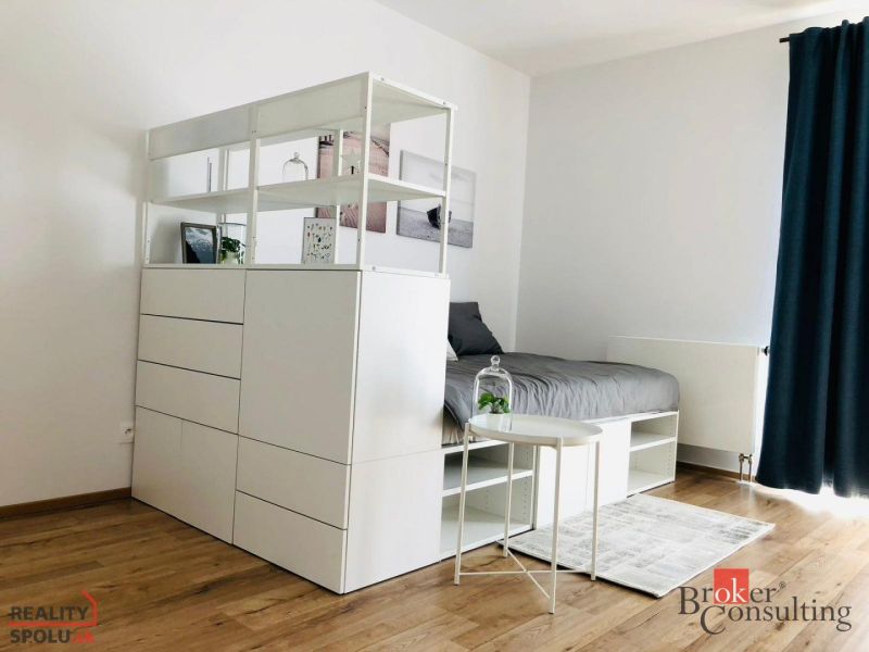 Ein Zimmer in einer Einzimmerwohnung mit einem Boden in Holzoptik, weißen Möbeln und einem Bett.