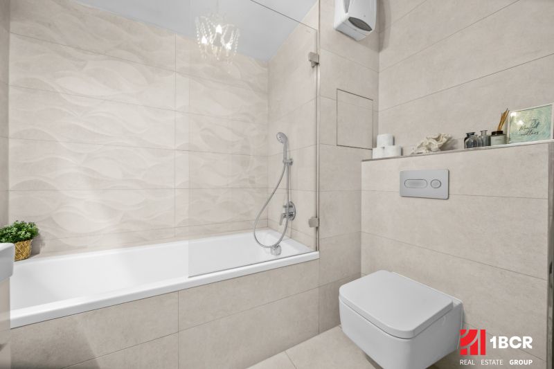 Badezimmer mit Badewanne und Toilette in einer 2-Zimmer-Wohnung mit heller Fliesenverkleidung.