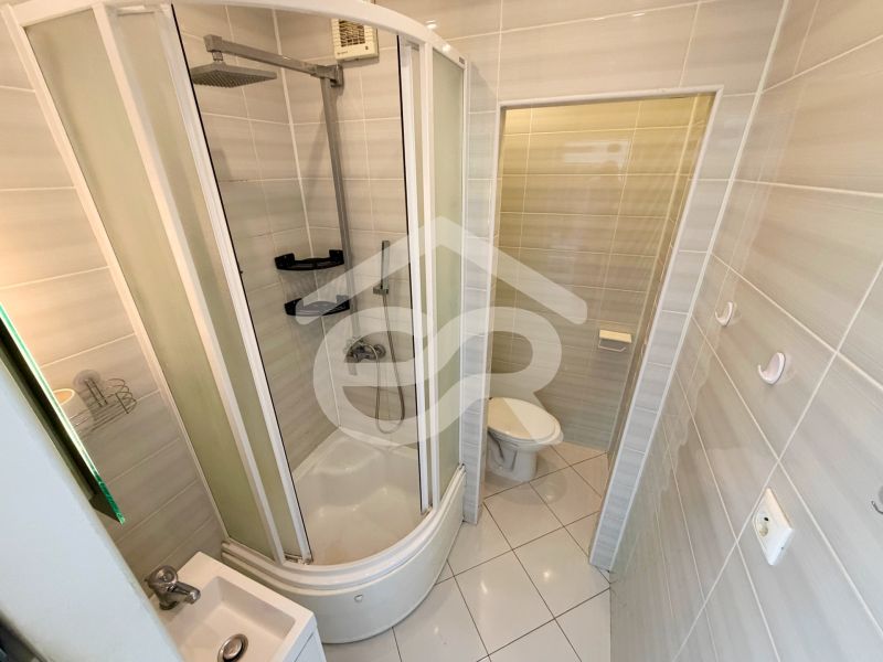 Badezimmer mit Duschkabine und Toilette in einer Einzimmerwohnung, helle Fliesen.