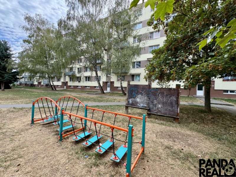 Fassade eines Wohnblocks in Topoľčany mit einem Kinderspielplatz und Vegetation rund um eine Einzimmerwohnung.