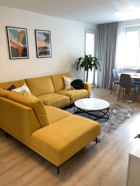Wohnzimmer einer 3-Zimmer-Wohnung mit großem gelbem Sofa, Holzboden und Pflanze.