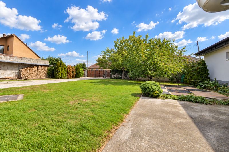 Ein Garten in Mosonmagyaróvár mit Rasen, Bäumen und einem Betonweg bei einem Einfamilienhaus.