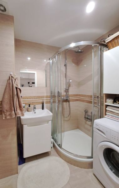 Badezimmer mit Waschbecken, Dusche und Waschmaschine in einer 3-Zimmer-Wohnung, beigefarbene Fliesen.