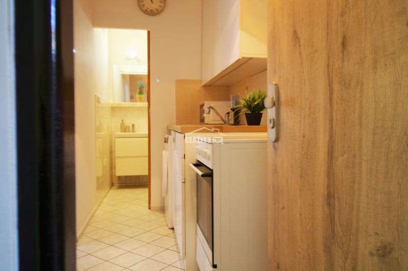 Kochnische in einem Einzimmerapartment mit weißen Schränken und Geräten, Fliesenboden.