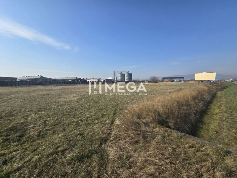 Gras und Produktionsräume in Hriadky unter klarem Himmel, mit einem Industriegebäude am Horizont.