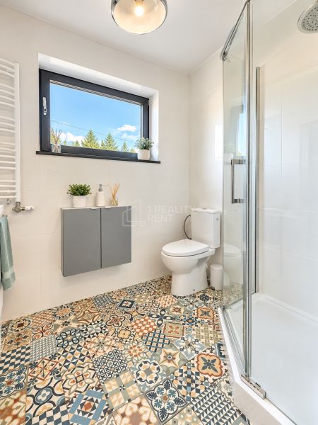 Badezimmer mit Dusche, Toilette und Fenster, bunte gemusterte Fliesen auf dem Boden.