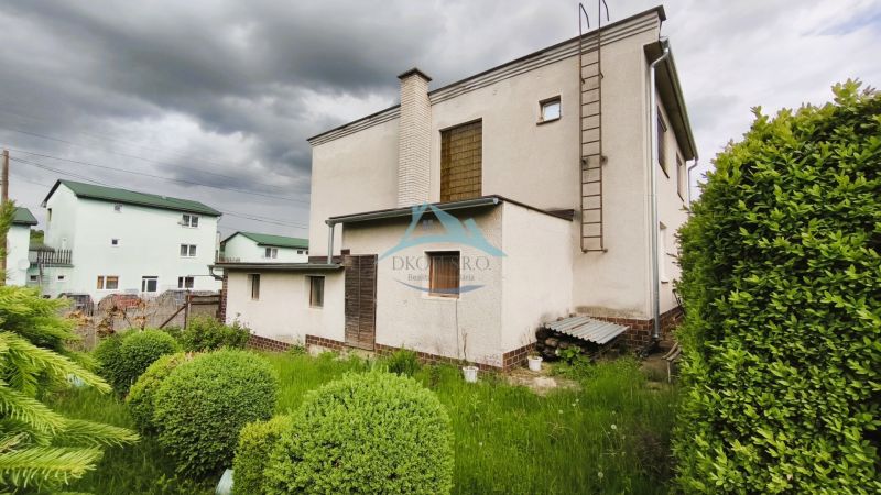 Ein Einfamilienhaus in Nenince mit Rasen und Sträuchern unter einem bewölkten Himmel.
