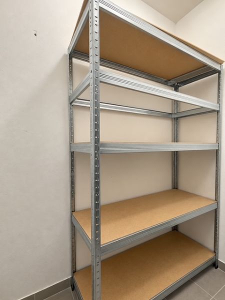 Ein Metallregal mit Regalböden in einer 2-Zimmer-Wohnung an einer weißen Wand und einem grauen Boden.