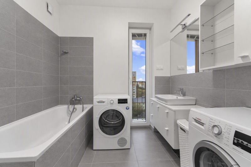 Badezimmer mit Badewanne, Waschmaschinen und Waschbecken in einer Zweizimmerwohnung. Graue Fliesen, großer Spiegel.