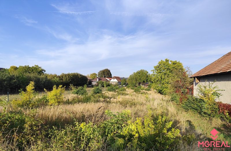 Grundstücke - Wohnen in Jasová, umgeben von Grün mit sichtbaren Gebäuden im Hintergrund.