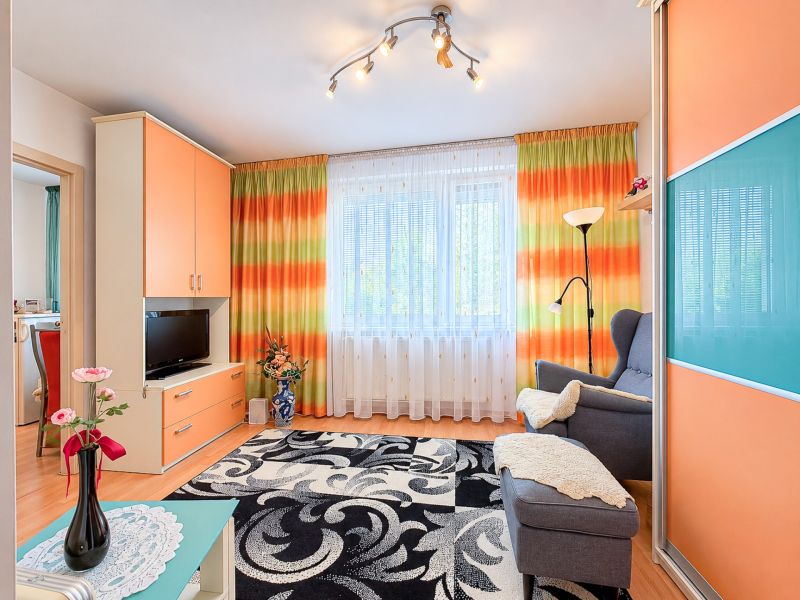 Wohnzimmer in einer Einzimmerwohnung mit bunten Vorhängen und einem schwarzen gemusterten Teppich.