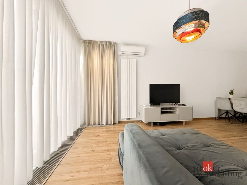 Wohnzimmer in einer Zweizimmerwohnung mit Sofa, Fernseher und Vorhängen auf einem Boden mit Holzdekor.