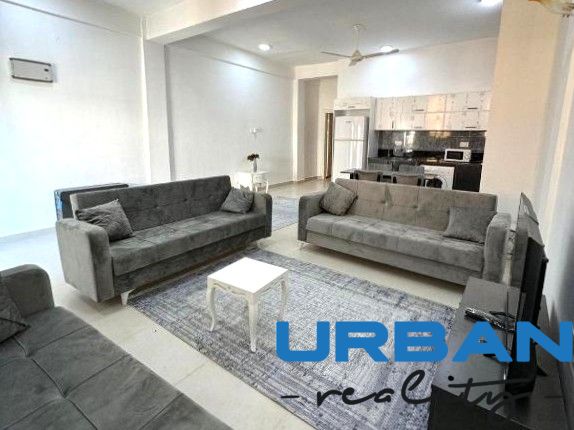 Wohnzimmer mit drei grauen Sofas, einem Couchtisch und einer Küche in einer 2-Zimmer-Wohnung.