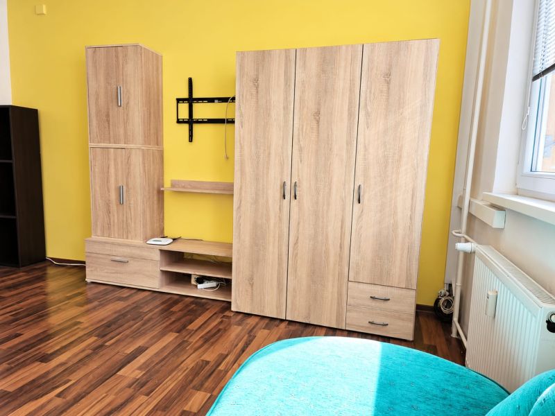 Einzimmerwohnung mit Holzboden, gelbe Wand, Schränke und TV-Halterung an der Wand.